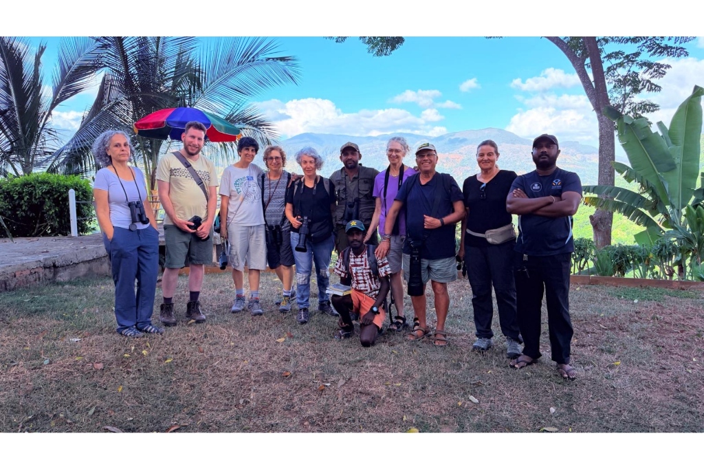 Group of international travelers and birdwatchers with binoculars and cameras smiling with guide Ravi against a scenic mountain backdrop