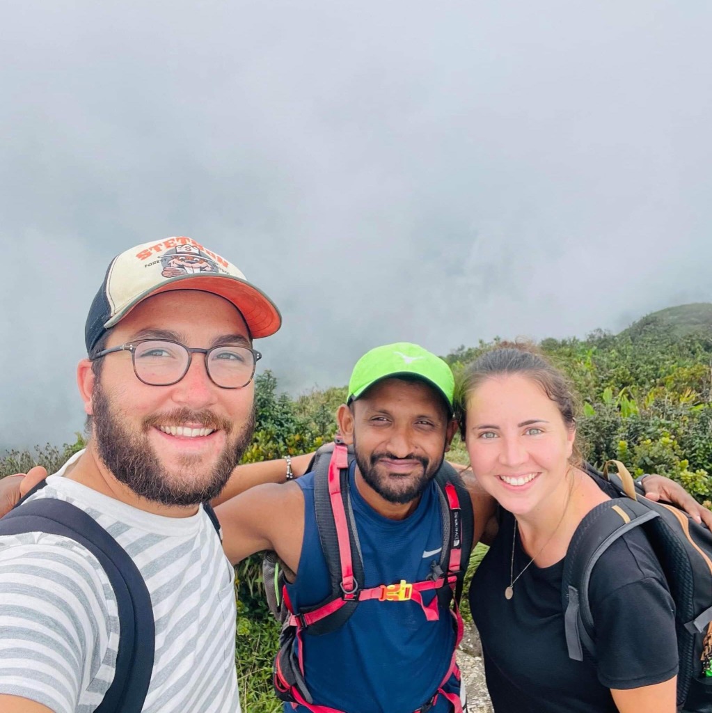 Guide Ravi and two backpackers smiling at a misty mountain summit viewpoint in the Knuckles Range