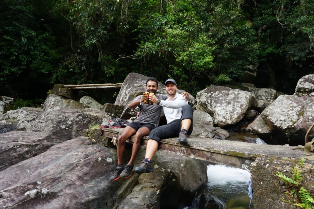 Guide Ravi and a guest trekker toasting drinks while relaxing on a bridge above a forest river cascade