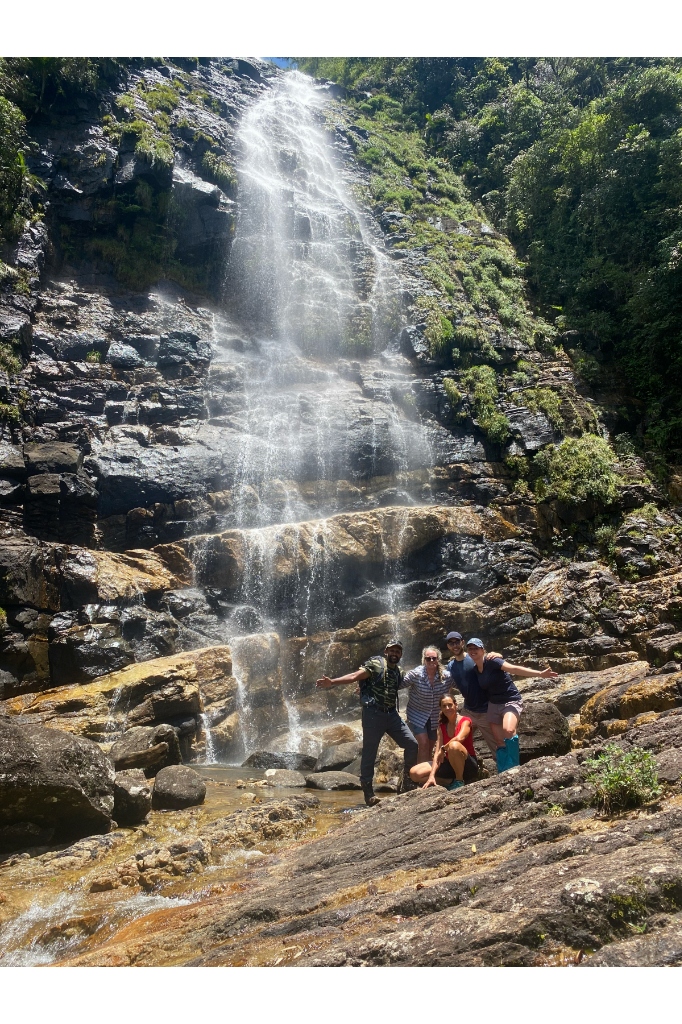 Adventure trekking group with guide Ravi celebrating with open arms beneath a towering Knuckles waterfall