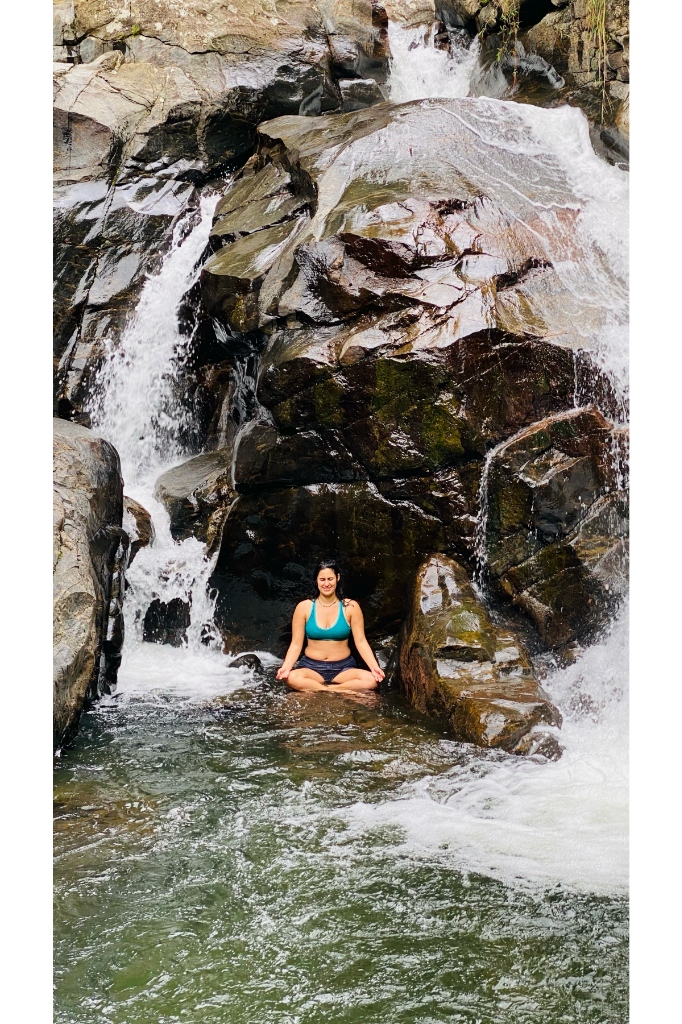 Traveler in teal swimwear in a serene meditation pose in a calm rock pool flanked by jungle waterfall cascades