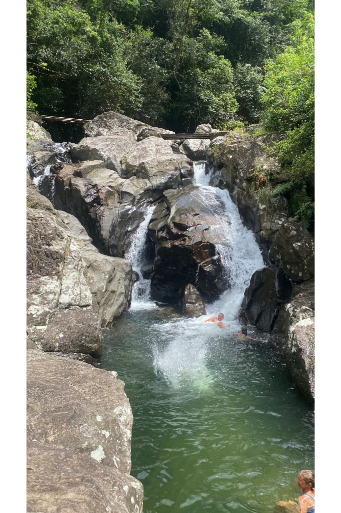 Adventure travelers swimming and jumping into a natural rock pool beneath a cascading waterfall in the High Knuckles