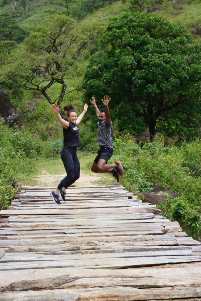 Two adventure hikers jumping mid-air in celebration on a wooden bridge along a forest trek